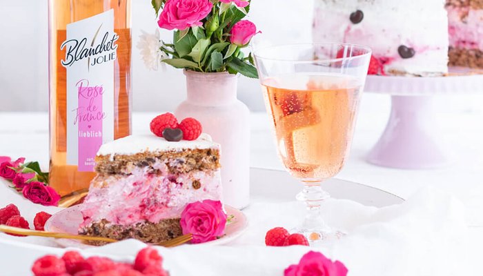 Ein Stück Torte mit einer rosa Füllung, umgeben von frischen Himbeeren und rosa Rosen, auf einem eleganten Tisch serviert.
