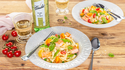 Ein Teller mit frischer Pasta, Lachs und buntem Gemüse, garniert mit Basilikum, auf einem rustikalen Holztisch.