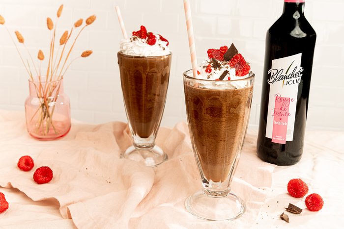 Zwei hohe Gläser mit Schokoladenmilchshake, dekoriert mit Sahne, Himbeeren und Schokoladenstückchen auf einem sanften Stoff.
