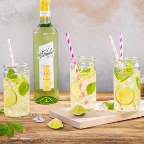 Drei Gläser mit erfrischenden Cocktails, garniert mit Limetten und frischem Basilikum, stehen auf einem Holzbrett.