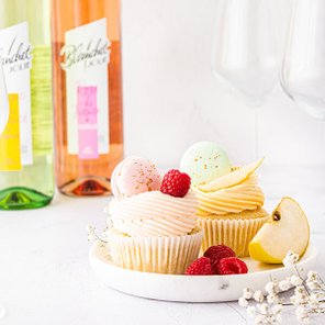 Eine Auswahl an bunten Cupcakes mit frischen Himbeeren und einer Apfelscheibe auf einem weißen Teller, im Hintergrund Weinflaschen.