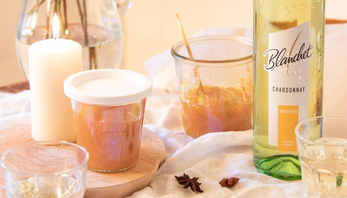 Ein Glas Chardonnay von Blanchet steht neben einem Glas mit einer goldenen Marmelade und einer brennenden Kerze auf einem Holzbrett.