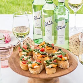 Ein rustikales Holzbrett präsentiert köstliche Bruschetta mit frischem Gemüse, umgeben von Gläsern Weißwein und Flaschen.