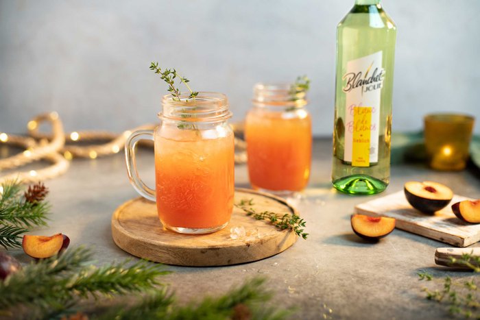 Zwei Gläser mit einem erfrischenden, orangefarbenen Getränk stehen auf einem Holzbrett, dekoriert mit Thymian und Pfirsichscheiben.