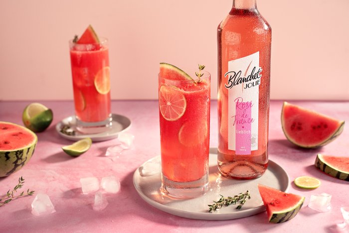 Zwei Gläser mit einem erfrischenden Wassermelonen-Drink stehen auf einem rosa Tisch, umgeben von frischen Früchten und Eiswürfeln.