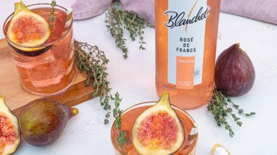 Ein Glas mit roséfarbenem Getränk, garniert mit einer Feige und frischen Kräutern, steht auf einem Holzbrett.