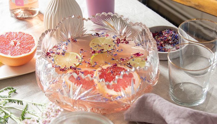 Eine elegante Glasschüssel mit fruchtigem Getränk, dekoriert mit Zitronenscheiben und essbaren Blüten, steht auf einem Tisch.