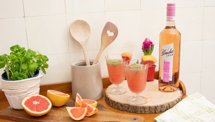 Ein rustikales Holzbrett zeigt zwei Cocktails, frische Grapefruits und eine Flasche Roséwein von Blanchet, umgeben von Küchenutensilien.