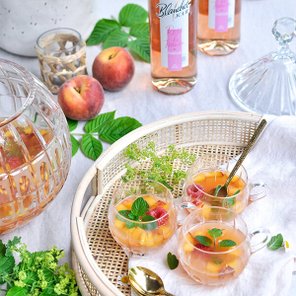 Ein geflochtener Korb enthält Gläser mit fruchtigem Getränk, dekoriert mit Minze und frischen Pfirsichen, umgeben von grünen Blättern.