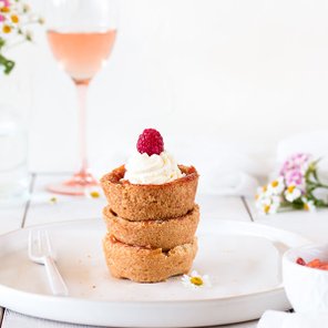Ein appetitlicher Dessertteller mit drei Schalen, gekrönt von Sahne und einer Himbeere, neben einem Glas Rosé und Blumen.
