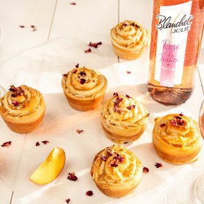 Sechs kunstvoll dekorierte Cupcakes mit Rosenblättern stehen auf einem weißen Tisch, umgeben von Gläsern mit Roséwein.
