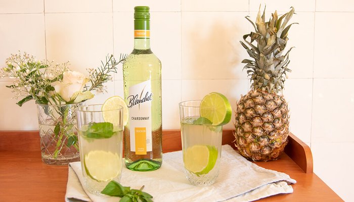 Ein Flasche Chardonnay von Blanchet steht neben zwei Gläsern mit Limetten und Minze auf einem Holztablett, umgeben von frischen Blumen.
