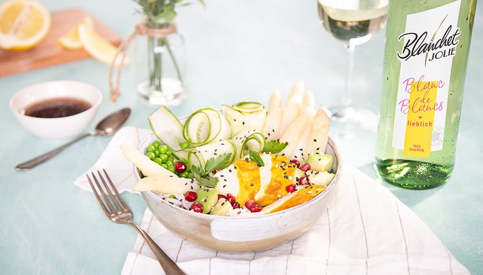 Eine kreative Salatkomposition mit frischen Zutaten, darunter Spargel und Gurken, neben einer Flasche Blanc de Blancs Wein.