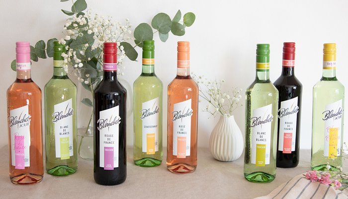 Sechs Flaschen Wein von Blanchet in verschiedenen Farben stehen auf einem Tisch, umgeben von frischen Blumen und grünen Blättern.