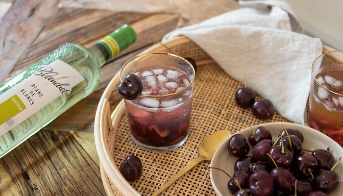 Eine Flasche Blanc de Blancs Wein liegt neben zwei Gläsern mit einem fruchtigen Getränk und frischen Kirschen.