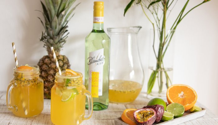 Zwei Gläser mit bunten Cocktails stehen vor einer Ananas und einer Flasche Blanchet, umgeben von frischen Früchten.