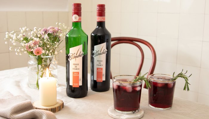 Zwei Flaschen Wein stehen neben einem Glas mit einem dunklen Getränk, dekoriert mit Rosmarin und Eiswürfeln.