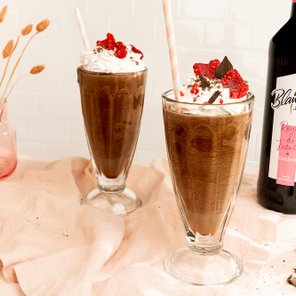 Zwei hohe Gläser mit Schokoladenmilchshake, dekoriert mit Sahne, Himbeeren und Schokoladenstückchen auf einem sanften Stoff.