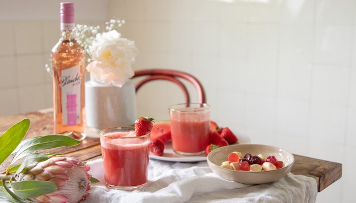 Zwei Gläser mit frischem Erdbeersaft stehen auf einem Holztisch, umgeben von frischen Früchten und einer Flasche Roséwein.