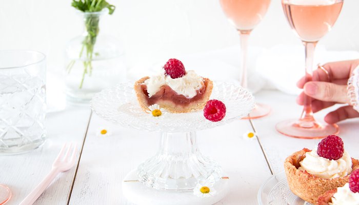 Ein zarter Dessertteller mit einer kleinen Torte, garniert mit Sahne und frischen Himbeeren, umgeben von Gläsern mit Rosé.
