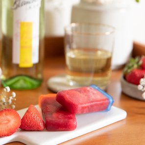 Auf einem Holztablett liegen rote Eis am Stiel neben frischen Erdbeeren und einem Glas Weißwein.