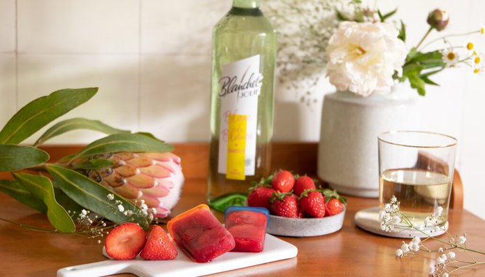 Ein rustikaler Holztisch mit frischen Erdbeeren, bunten Eiswürfeln und einer Flasche Likör im Hintergrund.