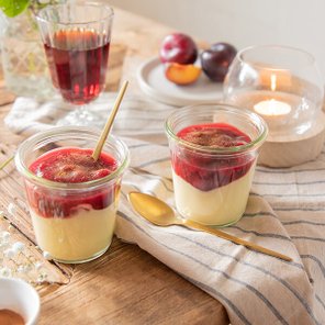Zwei Gläser mit Pudding, bedeckt mit roter Fruchtsauce, stehen auf einem Holztisch neben einer brennenden Kerze.