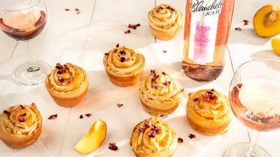 Sechs kunstvoll dekorierte Cupcakes mit Rosenblättern stehen auf einem weißen Tisch, umgeben von Gläsern mit Roséwein.