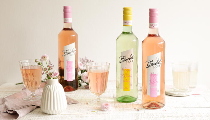 Drei Flaschen Wein von Blanchet stehen auf einem Tisch, umgeben von Gläsern und zarten Blumen, die eine fröhliche Atmosphäre schaffen.