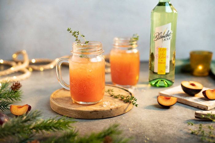 Zwei Gläser mit einem erfrischenden, orangefarbenen Getränk stehen auf einem Holzbrett, dekoriert mit Thymian und Pfirsichscheiben.