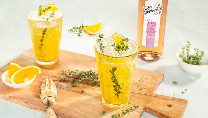 Zwei Gläser mit einem erfrischenden Getränk, garniert mit Thymian und einer Orangenscheibe, stehen auf einem Holzbrett.