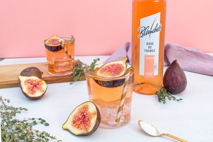 Ein erfrischendes Glas Roséwein mit einer Feige und einem Strohhalm, umgeben von frischen Kräutern auf einem hellen Tisch.