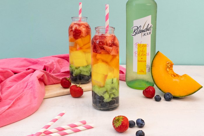 Zwei Flaschen mit buntem Fruchtcocktail stehen auf einem Tisch, umgeben von frischen Beeren und einer Melone.