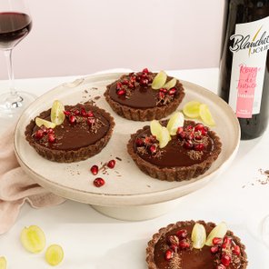 Drei köstliche Schokoladentörtchen mit Granatapfelkernen und Trauben auf einem hellen Teller, umgeben von einem weichen Tuch.