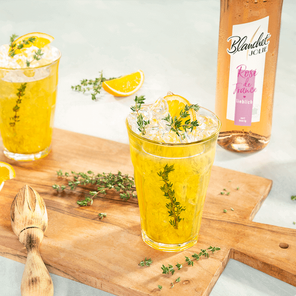 Zwei Gläser mit einem erfrischenden gelben Getränk, garniert mit Thymian und einer Orangenscheibe, auf einem Holzbrett.