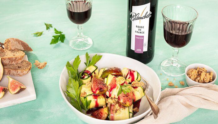 Ein köstlicher Salat mit frischen Feigen und Zucchini, serviert mit Rotwein und knusprigem Brot auf einem hellen Tisch.