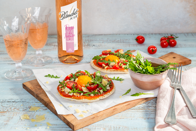 Zwei köstliche Flammkuchen mit frischen Tomaten, Rucola und einem Eigelb, serviert auf einem Holzbrett.