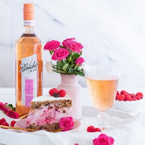 Ein eleganter Tisch mit einer Flasche Roséwein, einem Glas, einer Torte und frischen Himbeeren, umgeben von rosa Blumen.