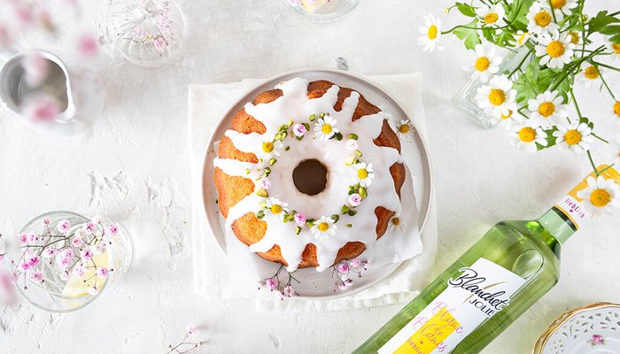 Ein saftiger Gugelhupf mit weißem Zuckerguss und essbaren Blumen dekoriert, umgeben von frischen Blumen und Gläsern.
