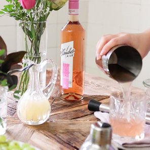 Eine Hand gießt einen Cocktail aus einem Shaker in ein Glas, während eine Flasche Roséwein im Hintergrund steht.