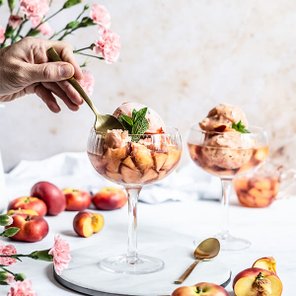Eine Hand mit einer goldenen Gabel schöpft fruchtiges Eis aus einem eleganten Glas mit Pfirsichstücken und Minze.
