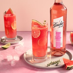 Zwei Gläser mit einem erfrischenden Wassermelonen-Drink stehen auf einem rosa Tisch, umgeben von frischen Früchten und Eiswürfeln.