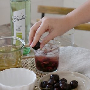 Eine Hand greift nach einer Kirsche, um sie in ein Glas mit Eis und einem dunklen Getränk zu geben.