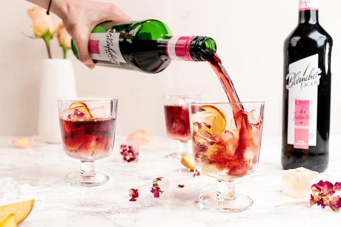 Ein Handgelenk gießt roten Wein in ein Glas mit Eis, umgeben von frischen Blütenblättern und einer Flasche im Hintergrund.