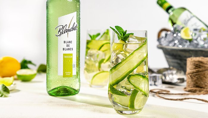 Auf einem hellen Tisch steht ein Glas mit einem kühlen Getränk, dekoriert mit Gurken und Minze, neben einer Flasche Wein.