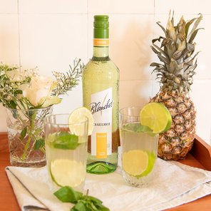 Ein Flasche Chardonnay von Blanchet steht zwischen zwei Gläsern mit Limetten und Minze, umgeben von einer Ananas und frischen Blumen.