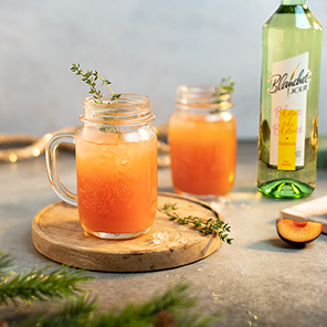 Zwei Gläser mit einem fruchtigen Getränk stehen auf einem Holzbrett, umgeben von frischen Kräutern und Pflaumenstücken.