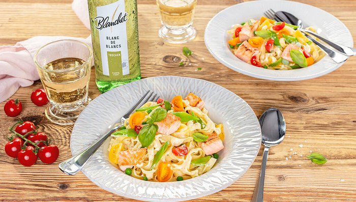Ein stilvoll angerichteter Pastateller mit Lachs und buntem Gemüse, begleitet von einem Glas Weißwein auf einem rustikalen Tisch.