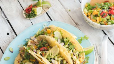 Ein Teller mit drei üppig gefüllten Tacos, garniert mit frischen Kräutern, Limetten und buntem Gemüse auf einem blauen Teller.