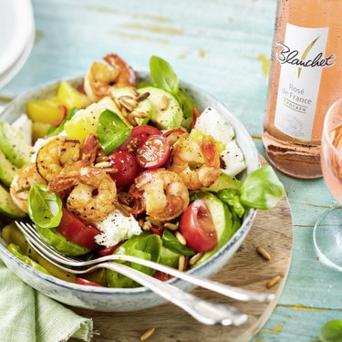 Ein bunter Salat mit Garnelen, Tomaten, Avocado und frischem Basilikum, serviert in einer Schüssel auf einem Holzbrett.